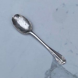 May include: A silver teaspoon with a rounded bowl and a long, slender handle. The spoon has a slightly worn, antique look.