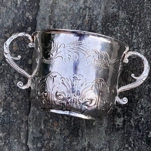 May include: A silver cup with two handles and an ornate design. The cup is sitting on a gray, textured surface.