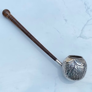 Puede incluir: Una tetera de plata con un mango de madera. La tetera tiene un diseño adornado con hojas y un borde de cuerda. El mango es de madera marrón oscuro.