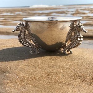 May include: A silver bowl with two seahorse handles. The bowl is sitting on a sandy beach.