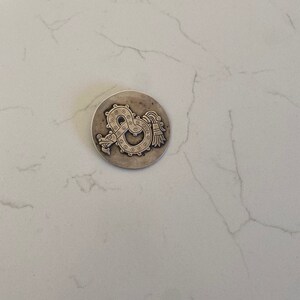 May include: A silver-toned pendant featuring a detailed design of a serpent with a feathered headdress. The serpent is curled into a circle with its tail in its mouth.