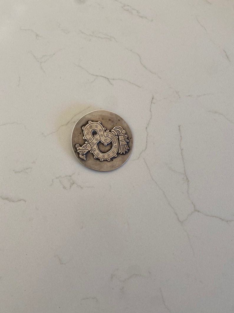 May include: A silver-toned pendant featuring a detailed design of a serpent with a feathered headdress. The serpent is curled into a circle with its tail in its mouth.
