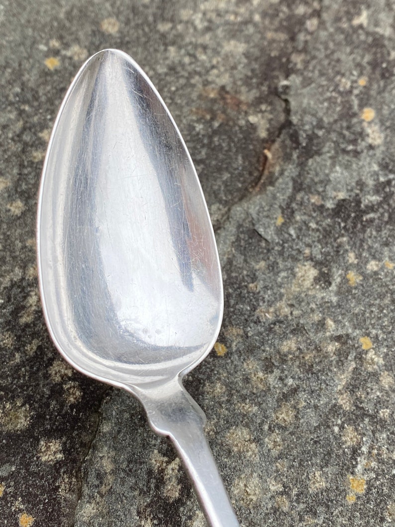 May include: A silver spoon with a rounded bowl and a long, slender handle. The spoon is lying on a rough, gray surface.