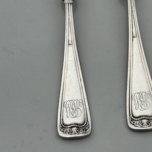 May include: Close-up of two silver spoons with ornate, leaf-like designs at the top and decorative flourishes at the base. Each spoon features an engraved monogram in a script font. The spoons are set against a gray background.