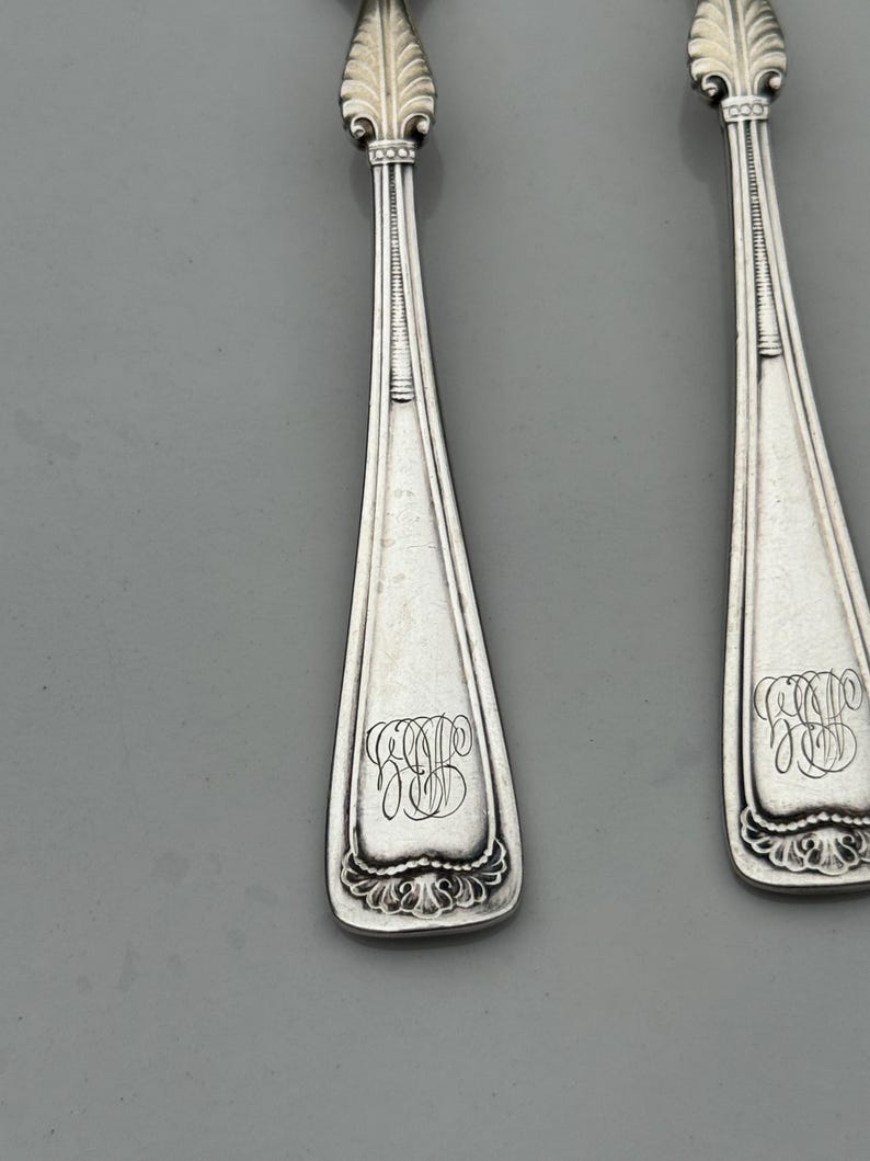 May include: Close-up of two silver spoons with ornate, leaf-like designs at the top and decorative flourishes at the base. Each spoon features an engraved monogram in a script font. The spoons are set against a gray background.