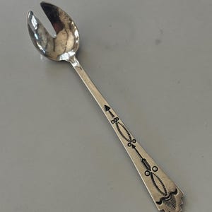 Peut inclure: Une fourchette argentée avec une tête fendue en forme de cuillère. Le manche est orné d'un motif linéaire de flèches, de cercles et de formes géométriques. Cet ustensile est probablement destiné à servir ou à manger.