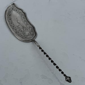 May include: A silver serving utensil with an ornate, curved blade and a twisted handle. The blade features an engraved floral design and a scalloped edge. The handle ends in a decorative, rounded finial.