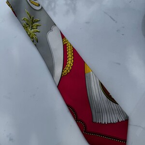 Puede incluir: Un pa&ntilde;uelo de seda con fondo gris y borde rojo. El pa&ntilde;uelo presenta dise&ntilde;os de cadenas y borlas doradas, junto con un dise&ntilde;o blanco y gris. El pa&ntilde;uelo mide aproximadamente 7.6 cm de ancho y 127 cm de largo.