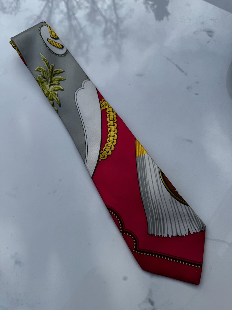 Puede incluir: Un pa&ntilde;uelo de seda con fondo gris y borde rojo. El pa&ntilde;uelo presenta dise&ntilde;os de cadenas y borlas doradas, junto con un dise&ntilde;o blanco y gris. El pa&ntilde;uelo mide aproximadamente 7.6 cm de ancho y 127 cm de largo.