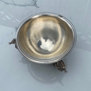 May include: A silver bowl with a round, smooth interior and three ornate, animal-shaped feet. The bowl is sitting on a white marble surface.