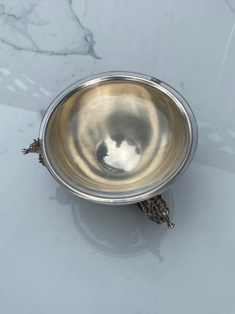 May include: A silver bowl with a round, smooth interior and three ornate, animal-shaped feet. The bowl is sitting on a white marble surface.