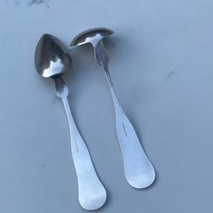 May include: Two silver spoons with ornate handles. The spoons have a rounded, oval-shaped bowl.