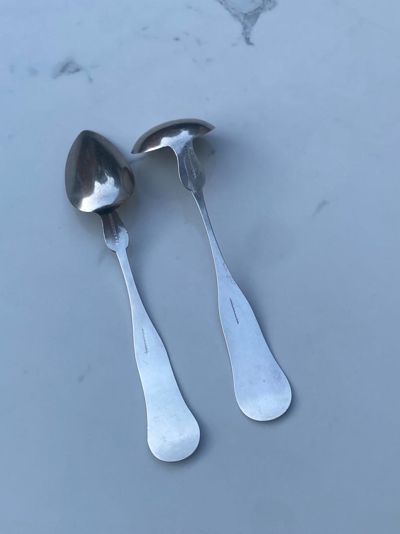 May include: Two silver spoons with ornate handles. The spoons have a rounded, oval-shaped bowl.