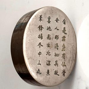 Puede incluir: Un recipiente redondo de color plateado con caracteres chinos negros grabados en la parte superior. El recipiente tiene una banda marrón oscuro alrededor del lateral. El texto está en columnas verticales.