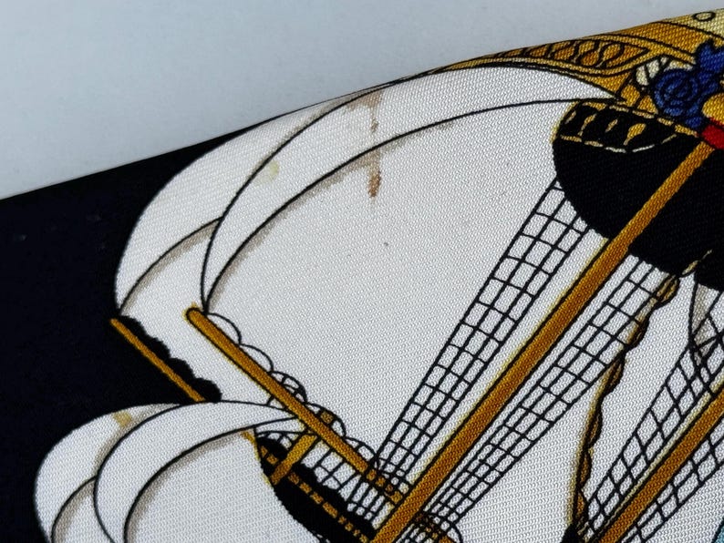 Puede incluir: Primer plano de un pa&ntilde;uelo de seda estampado con un dise&ntilde;o de velero. El pa&ntilde;uelo tiene un fondo blanco con detalles en negro y dorado. Las velas del barco son blancas y el aparejo es negro.