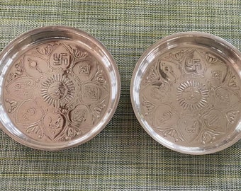 Può includere: Due piatti d'argento con disegni intricati. I piatti presentano un sole con un viso, una svastica e altri simboli. I piatti sono su una superficie verde testurizzata.