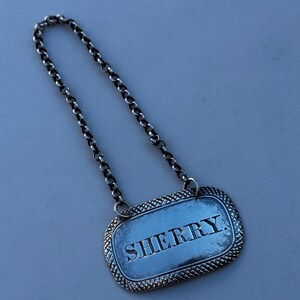 May include: A silver-toned rectangular wine label with the word "SHERRY" engraved in capital letters. The label has a decorative border and is attached to a chain. The chain is made of small, interlocking links.