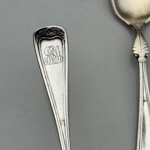 May include: Close-up of two antique silver spoons. One spoon has a rectangular handle with an ornate floral design at the top and engraved initials. The other spoon has a rounded bowl and a decorative handle. Both spoons are on a gray background.