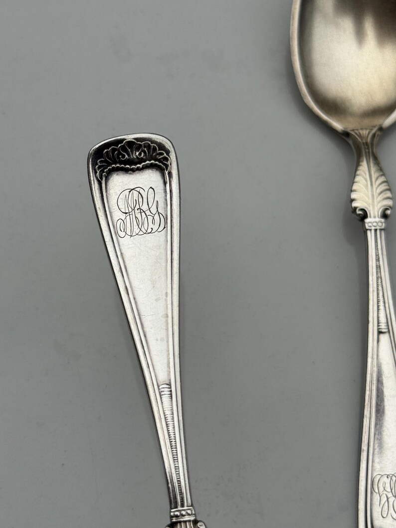 May include: Close-up of two antique silver spoons. One spoon has a rectangular handle with an ornate floral design at the top and engraved initials. The other spoon has a rounded bowl and a decorative handle. Both spoons are on a gray background.