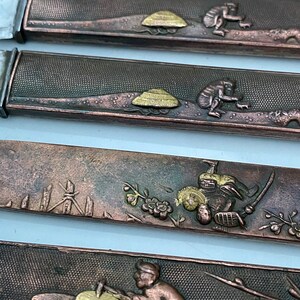 K&ouml;nnte beinhalten: Nahaufnahme von vier verzierten japanischen Schwertklingen, oder Tsuba, mit aufwendigen Designs in Gold und Kupfer. Die Designs beinhalten einen Pilz, einen Affen, einen Samurai und eine Person, die Holz schnitzt.