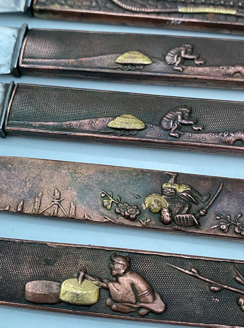 K&ouml;nnte beinhalten: Nahaufnahme von vier verzierten japanischen Schwertklingen, oder Tsuba, mit aufwendigen Designs in Gold und Kupfer. Die Designs beinhalten einen Pilz, einen Affen, einen Samurai und eine Person, die Holz schnitzt.