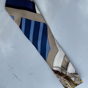 Puede incluir: Un pa&ntilde;uelo de seda con tem&aacute;tica ecuestre. El pa&ntilde;uelo presenta un patr&oacute;n de azul, beige y blanco con un dise&ntilde;o de aparejos de caballo y rayas. El pa&ntilde;uelo es largo y estrecho.
