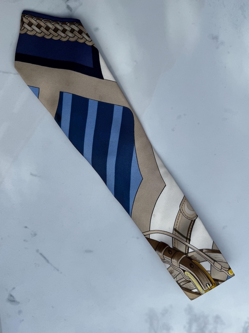 Puede incluir: Un pa&ntilde;uelo de seda con tem&aacute;tica ecuestre. El pa&ntilde;uelo presenta un patr&oacute;n de azul, beige y blanco con un dise&ntilde;o de aparejos de caballo y rayas. El pa&ntilde;uelo es largo y estrecho.