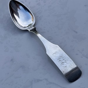 May include: A silver-toned metal spoon with a polished, reflective oval bowl and a flat, wide handle. The handle features an ornate, cursive monogram engraving of the letters M, C, and B, with a slightly darkened, tarnished finish at the very end.