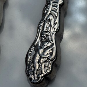 May include: Close-up of a silver utensil handle with detailed relief carvings. The design features figures and floral patterns, creating a vintage aesthetic. The handle has a curved shape and a polished finish.