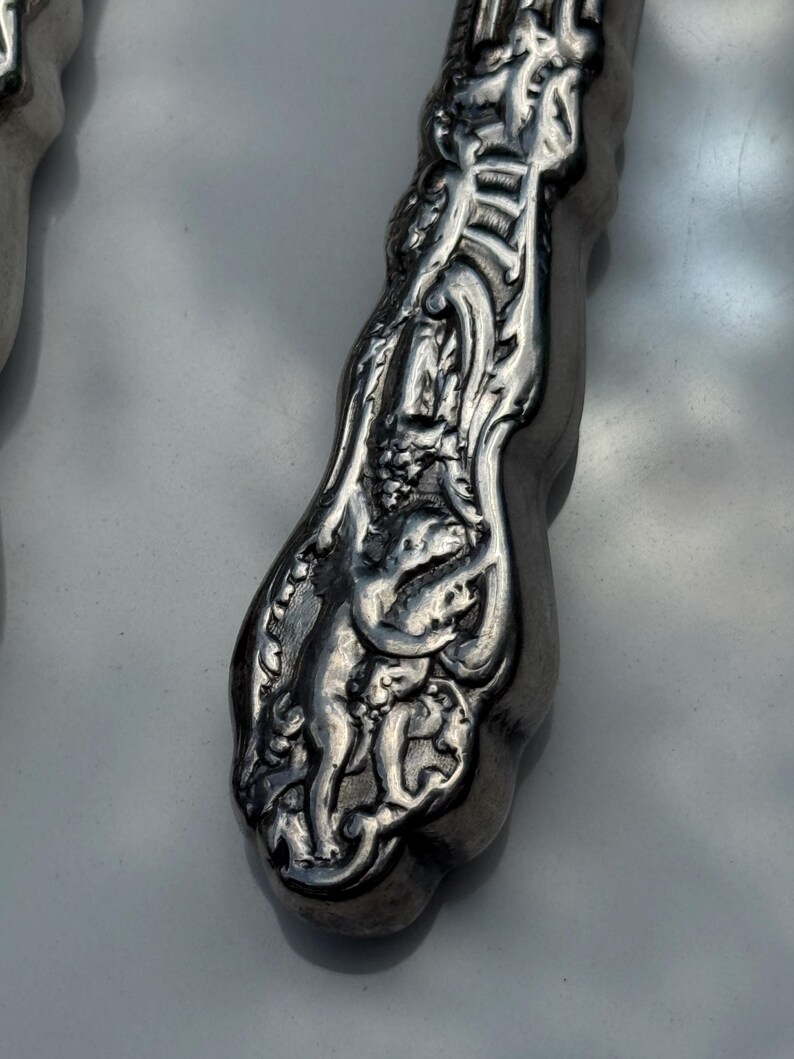 May include: Close-up of a silver utensil handle with detailed relief carvings. The design features figures and floral patterns, creating a vintage aesthetic. The handle has a curved shape and a polished finish.