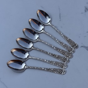 May include: A set of six silver-toned metal spoons arranged in a fan pattern. Each spoon features a polished oval bowl and a handle decorated with a detailed, raised floral relief pattern that extends from the neck to the rounded end of the handle.