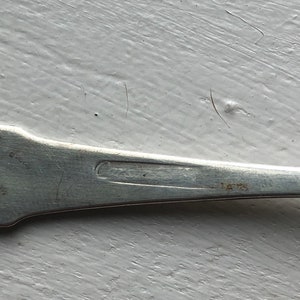 Puede incluir: Primer plano de la parte inferior de una cuchara de plata con una inscripci&oacute;n tenue.