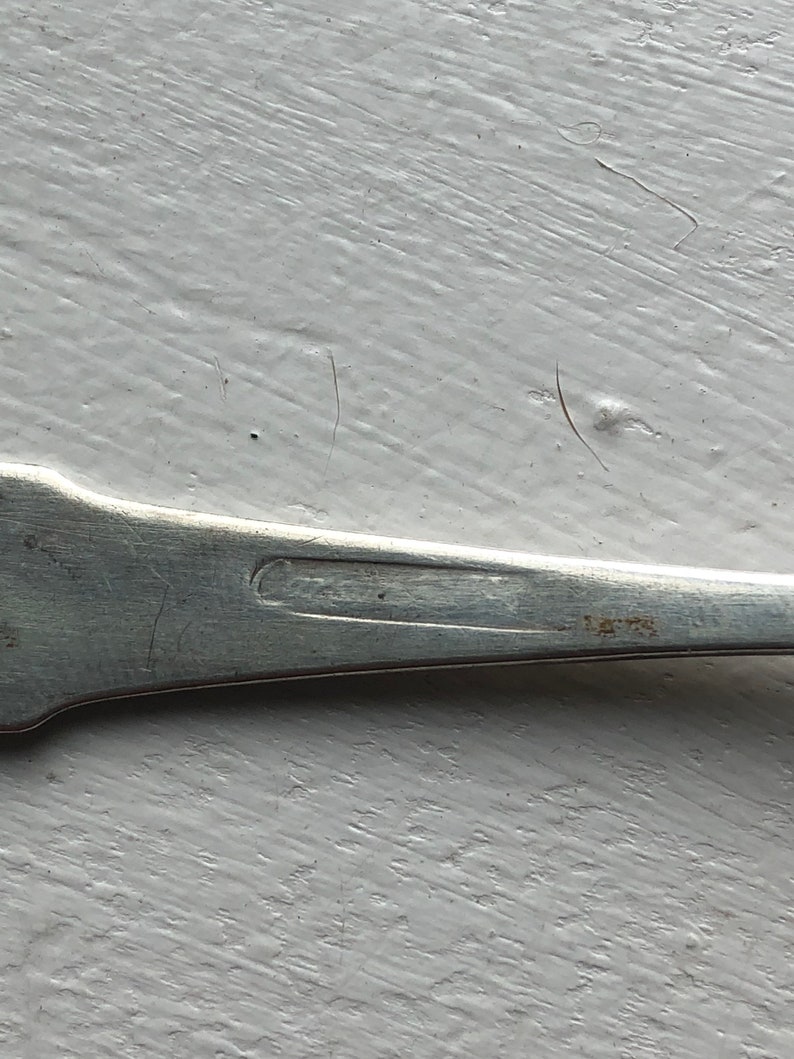 Puede incluir: Primer plano de la parte inferior de una cuchara de plata con una inscripci&oacute;n tenue.