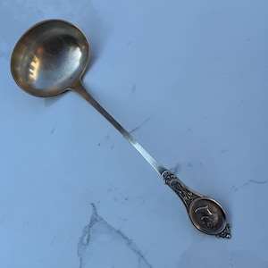 May include: A silver ladle with a round bowl and a long, slender handle. The handle features an ornate design with a circular emblem at the end. The ladle is likely vintage or antique, and is suitable for serving soups and sauces.