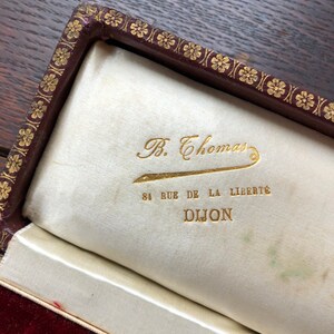 May include: A close-up of a burgundy velvet jewelry box with a gold embossed floral design. The interior is lined with white satin and features a gold inscription that reads "B. Thomas 84 Rue de la Liberte Dijon".