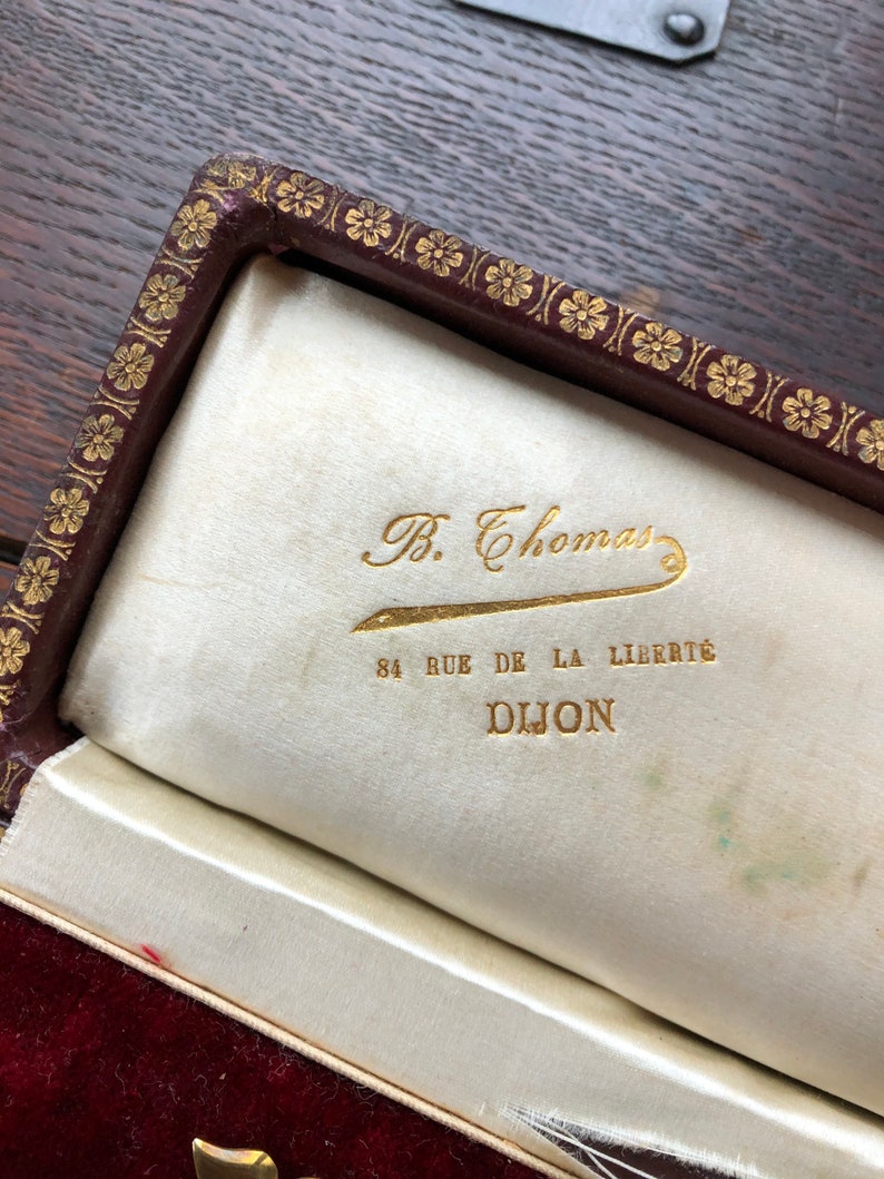 May include: A close-up of a burgundy velvet jewelry box with a gold embossed floral design. The interior is lined with white satin and features a gold inscription that reads "B. Thomas 84 Rue de la Liberte Dijon".