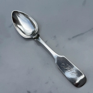 May include: A silver spoon with a rounded bowl and a long, slender handle. The handle has an engraved monogram. The spoon is set against a light gray background.