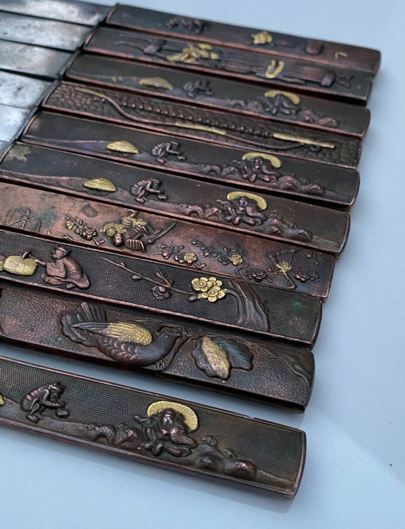 K&ouml;nnte beinhalten: Ein Satz von 10 japanischen Messern mit aufwendigen Designs auf den Griffen. Die Griffe sind aus dunklem Metall mit goldenen Akzenten. Die Designs beinhalten V&ouml;gel, Blumen und andere traditionelle japanische Motive.