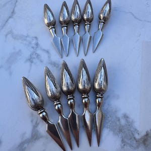 May include: A set of ten silver corn holders, each featuring a textured, ear-of-corn shaped handle with the word STERLING stamped on the side. The holders have a pointed, metallic prong end designed for securing corn on the cob during consumption.