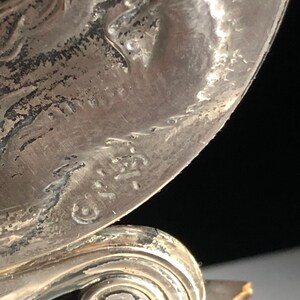 May include: Close-up of a silver-toned metal object with a swirl design at the base. The object has a raised inscription that reads "G.I.R.A. 1921".