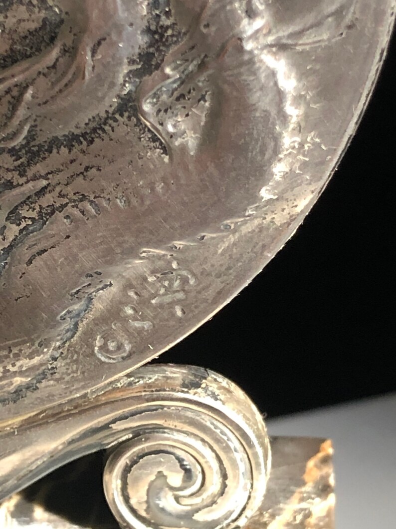 May include: Close-up of a silver-toned metal object with a swirl design at the base. The object has a raised inscription that reads "G.I.R.A. 1921".