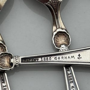 May include: Close-up of antique silver spoons. The spoons have a shell design on the handle and the text "PATENT 1880 GORHAM" is engraved on the spoon. The spoons are arranged on a light gray surface.