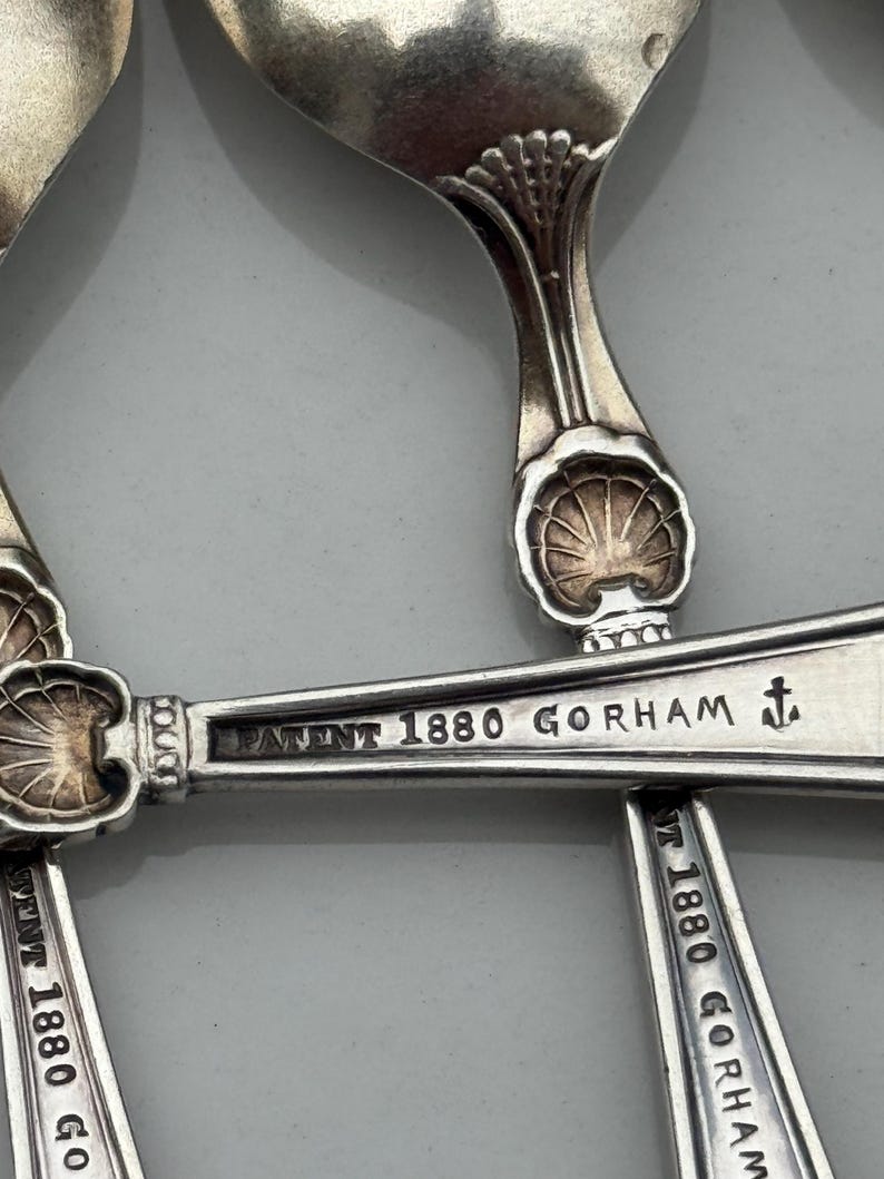 May include: Close-up of antique silver spoons. The spoons have a shell design on the handle and the text "PATENT 1880 GORHAM" is engraved on the spoon. The spoons are arranged on a light gray surface.