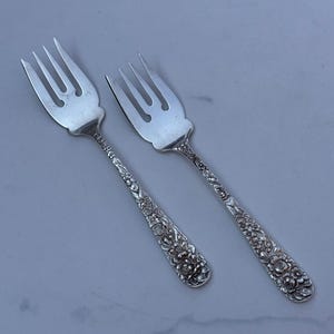 May include: Two silver-toned metal forks featuring four tines and a wide, rounded base. The handles are decorated with an intricate, raised floral pattern that extends from the base of the tines down to the rounded ends of the handles.