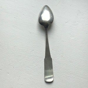 Puede incluir: Una cucharadita de plata con un cuenco redondeado de forma ovalada y un mango simple y recto.