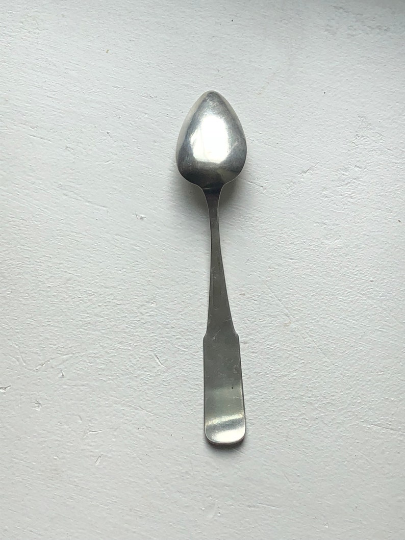 Puede incluir: Una cucharadita de plata con un cuenco redondeado de forma ovalada y un mango simple y recto.