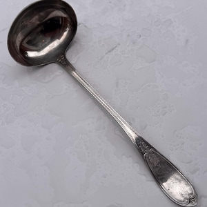 May include: A silver-toned metal soup ladle featuring a deep, rounded bowl and a long, slender handle. The handle is decorated with intricate, embossed scrollwork patterns at the base and near the bowl, showcasing a polished, reflective finish.