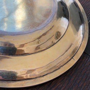 Puede incluir: La parte inferior de una fuente redonda de servir de lat&oacute;n con un borde ligeramente elevado. La fuente tiene una superficie lisa y pulida con algunos ara&ntilde;azos.