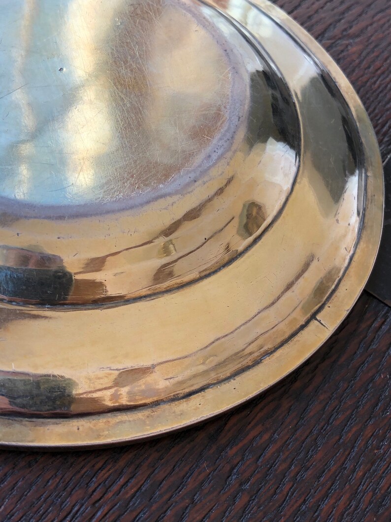 Puede incluir: La parte inferior de una fuente redonda de servir de lat&oacute;n con un borde ligeramente elevado. La fuente tiene una superficie lisa y pulida con algunos ara&ntilde;azos.