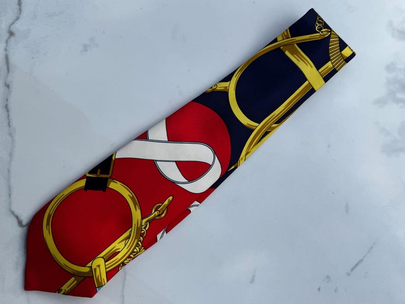 Puede incluir: Corbata de seda con dise&ntilde;o ecuestre rojo, azul marino y dorado. La corbata presenta un patr&oacute;n repetido de bocados de caballo dorados, cadenas y un dise&ntilde;o de cinta blanca sobre un fondo rojo y azul marino.